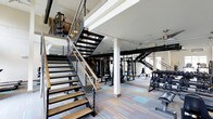 Located at 1701 Atria Ct in Raleigh, NC, this two-story fitness center offers a sleek and contemporary workout environment designed to accommodate a wide range of exercise needs. The space is anchored by a striking open-tread staircase with dark metal framing and wood-finished steps, accented by cable railings that contribute to the facility's modern industrial aesthetic.

The main floor features a spacious, open layout with durable multi-tone plank flooring in cool gray and blue hues. It is outfitted with a comprehensive selection of strength training equipment, including a power rack, cable machines, and an extensive dumbbell rack, alongside cardio stations. Structural columns are integrated into the design without interrupting the flow of the space.

The upper level, visible from the main floor through the open mezzanine, continues the fitness programming with additional equipment, making full use of the vertical space. Track lighting throughout both floors ensures excellent illumination across all workout zones.

High ceilings and an open mezzanine design create an airy, expansive atmosphere, while the consistent use of dark metal accents and neutral tones gives the facility a cohesive, professional look. This fitness center is thoughtfully designed to serve residents with both functionality and style.