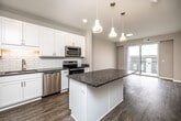 This efficient yet thoughtfully designed 1-bedroom, 1-bathroom unit offers a cohesive modern aesthetic across a single floor. The open-concept kitchen and living area serves as the heart of the space, where the kitchen stands out with white shaker cabinetry, a subway tile backsplash, dark granite countertops, stainless steel appliances, and a central island with pendant lighting overhead — ideal for both cooking and casual dining. The living area extends naturally from the kitchen and opens to a private balcony through sliding glass doors, allowing for an abundance of natural light and outdoor views. Consistent wood-look flooring runs throughout the main living areas, complementing the neutral warm-toned walls. The bedroom features plush carpet, a window with natural light, and access to a walk-in closet with built-in shelving. The full bathroom includes a white vanity with dark granite countertops, a large mirror, wood-look flooring, and accessible grab bars. A dedicated laundry area adds everyday convenience. The overall ambiance is clean, contemporary, and functional, with cohesive finishes tying the entire space together seamlessly.