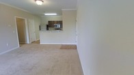 This single-story space showcases an open-concept design that seamlessly connects the living area to the kitchen. The living area is finished with neutral-toned wall-to-wall carpeting, creating a comfortable and cohesive atmosphere throughout. Crown molding lines the ceilings, adding a refined architectural detail that enhances the overall aesthetic.

The kitchen is positioned at the rear of the open layout, separated from the living space by a raised breakfast bar that provides both a casual dining option and a natural visual divider. Dark wood cabinetry contrasts with the light-colored walls, giving the kitchen a grounded, classic look. Stainless steel appliances, including a refrigerator, microwave, and range, are integrated into the space, offering modern functionality.

The transition from carpeted living area to hard flooring in the kitchen adds textural variety underfoot. Recessed and overhead lighting fixtures illuminate the space evenly. The light, neutral color palette applied throughout the walls and flooring contributes to a clean, airy ambiance, making the space feel open and easy to personalize.