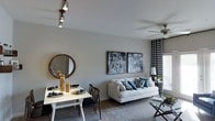 This single-floor space offers a well-designed open-concept layout that integrates the dining and living areas into one cohesive, inviting environment. The living area is anchored by a plush white sofa accented with patterned throw pillows in navy and teal, complemented by a sleek glass coffee table. Bold typographic wall art serves as a striking focal point, adding a modern, urban character to the space. A ceiling fan with an integrated light fixture enhances both comfort and functionality overhead. The dining area features a clean white table set for four, with dark wood chairs and elegantly staged place settings in navy and gold. A round wood-framed mirror on the wall adds depth and visual interest to the dining space. Track lighting mounted on the ceiling provides targeted illumination, highlighting key areas throughout. Neutral gray walls and coordinating carpet flooring create a calm, cohesive backdrop, while chevron-patterned curtains frame the windows and introduce a dynamic textural element. Natural light filters in through the covered windows, contributing to the bright and welcoming ambiance of the overall space.