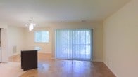 This single-floor space features an open-concept layout that seamlessly connects the living and dining areas. The living area is highlighted by large sliding glass doors dressed with vertical blinds, allowing generous natural light to fill the room while providing access to the outdoors. A side window further enhances the brightness of the space, offering views of surrounding greenery. The flooring throughout is warm-toned hardwood, adding a sense of continuity and warmth to the interior. The walls are finished in a neutral beige tone, creating a clean and versatile backdrop. The dining area, partially visible to one side, features a dark-toned countertop or bar surface and is illuminated by a pendant light fixture overhead, providing a subtle contrast to the lighter tones of the rest of the space. Ceiling-mounted HVAC vents are evenly distributed, ensuring consistent climate control throughout. The overall ambiance is bright, airy, and functional, with an understated, neutral aesthetic that lends itself well to a variety of interior design styles.