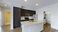 Located at 3443 Park Street in Grove City, Ohio, this single-floor space offers a clean, contemporary design with thoughtful finishes throughout. The kitchen serves as a standout feature, anchored by a spacious island topped with light granite countertops and an integrated double sink. Dark espresso-toned cabinetry extends floor to ceiling, providing generous storage while creating a striking visual contrast against the bright white walls. Stainless steel appliances — including a range with microwave above and a full-size refrigerator — complete the modern kitchen aesthetic. Recessed lighting illuminates the space efficiently, enhancing the open, airy feel. The flooring is a warm wood-tone plank, adding texture and continuity throughout the living areas. The overall design balances functionality with a modern sensibility, featuring neutral tones and clean lines that create a versatile and welcoming environment. The layout flows naturally, making the space feel open and well-organized. In-unit laundry connections are also visible, adding a practical convenience to this well-appointed single-floor property.