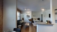 This single-floor studio is designed with an open-concept layout that efficiently combines living, sleeping, and dining functions within one cohesive space. The living area features a sleek dark sofa paired with a low-profile wooden coffee table and an accent butterfly chair, creating a comfortable and contemporary seating arrangement. Dark hardwood floors run throughout, anchoring the space with warmth and contrast against the light-toned walls.

The sleeping area is positioned toward the rear, furnished with a platform bed and complemented by decorative wall mirrors that add depth and visual interest. A wall-mounted mini-split air conditioning unit ensures year-round climate control. Track lighting along the ceiling provides adjustable, focused illumination, while a pendant light fixture adds a stylish touch near the kitchen counter.

A partial kitchen counter with granite-style surfacing is visible on one side, hinting at a dedicated cooking space. Windows along one wall admit natural light and offer city views, enhancing the urban character of the unit. The overall ambiance is modern and well-appointed, with thoughtful décor choices that maximize both style and functionality in a compact footprint.