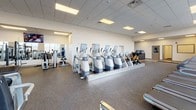 This modern fitness facility is thoughtfully organized across a single floor, offering a comprehensive range of workout zones to accommodate diverse training needs. Guests are welcomed at a clean, contemporary reception desk finished in teal and natural wood tones, with tile flooring that transitions seamlessly into rubber sport flooring throughout the main workout areas.

The expansive cardio zone is a standout feature, lined with rows of treadmills, ellipticals, stair climbers, and rowing machines positioned directly in front of tall, floor-to-ceiling windows. These large-pane windows flood the space with natural light and offer open views of the surrounding landscape, creating an energizing environment for cardio training.

The free weights area is well-stocked with a full rack of dumbbells, adjustable benches, and cable machines, complemented by full-wall mirrors that enhance the sense of space and allow users to monitor their form. Nearby, a functional training corner provides kettlebells, resistance bands, medicine balls, and open floor space for dynamic movement.

A dedicated indoor cycling studio houses a substantial fleet of stationary bikes arranged in rows, with mirrored walls and recessed lighting creating an immersive class environment. An additional multipurpose studio offers open floor space alongside stability balls, step platforms, yoga mats, and other group fitness accessories. Durable speckled rubber flooring throughout ensures safety and durability across all zones.