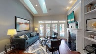This well-appointed clubhouse offers a thoughtfully designed single-floor layout that balances comfort and functionality. The main lounge area greets visitors with soaring vaulted ceilings, rich hardwood floors, and an abundance of natural light streaming through large French doors fitted with transom windows. A classic white-mantled fireplace serves as a focal point, complemented by a wall-mounted flat-screen television, stylish seating arrangements, and curated artwork. A striking tray ceiling with a skylight overhead adds architectural interest and further brightens the space.

A separate reception area features additional vaulted ceilings with crown molding, a ceiling fan, and plush tufted seating arranged on the hardwood floors. Gray accent walls display framed artwork and a console table with decorative accents, creating a refined yet approachable atmosphere.

The kitchen is equipped with white cabinetry, black appliances, and a pass-through bar counter with modern wire barstools — ideal for casual gatherings and community events. Recessed lighting throughout ensures the space is consistently well-lit. The overall ambiance is warm and inviting, with a polished aesthetic that makes the clubhouse a versatile hub for residents to gather, relax, and connect.