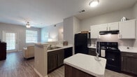 Located at 10127 State Highway 16 S in San Antonio, TX, this single-floor residence offers a thoughtfully designed layout with a seamless flow between living and dining spaces. The open-concept kitchen is a standout feature, equipped with black appliances including a refrigerator, electric range, and dishwasher, complemented by white upper cabinetry, dark lower cabinetry, and light granite-style countertops. A central kitchen island provides additional prep space and casual seating potential. Wood-look flooring runs throughout the unit, adding warmth and continuity across all rooms. The living area is bright and welcoming, with a large window that draws in natural light and offers outdoor views. Multiple bedrooms are present, each featuring ceiling fans, windows with blinds, and the same durable wood-look flooring. The primary bedroom includes an en-suite bathroom outfitted with a tiled tub-shower combination, white cabinetry, and classic fixtures. The overall color palette of soft grays and white creates a clean, neutral ambiance throughout the space. The functional floor plan, evident from the dollhouse view, makes efficient use of space while maintaining a comfortable and livable atmosphere.