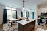 This contemporary single-floor residence is thoughtfully designed with a clean, modern aesthetic and an efficient layout that promotes comfort and connectivity throughout. A wide entry hallway with rich hardwood-style flooring leads into the heart of the home, where an open-concept kitchen, dining, and living area takes center stage. The kitchen is outfitted with two-tone dark wood and white cabinetry, quartz countertops, a glass tile backsplash, stainless steel appliances, and a central island with pendant lighting above — ideal for both everyday living and entertaining. The adjacent dining and living areas are bathed in natural light from expansive windows dressed with teal drapes, offering pleasant outdoor views. The living room features corner windows that further amplify the airy, light-filled atmosphere. Two well-proportioned bedrooms provide comfortable retreats, each with soft carpeting and large windows. The primary bedroom is served by an en-suite bathroom complete with a walk-in glass shower, dark wood vanity, and built-in shelving. A second full bathroom mirrors the same refined finishes with wood-look flooring and a sleek vanity. Consistent use of neutral tones, modern fixtures, and quality materials throughout creates a cohesive and polished living environment.