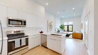 This contemporary single-floor residence showcases a clean, modern design characterized by an all-white palette, light hardwood flooring, and an abundance of natural light throughout. The open-concept kitchen flows seamlessly into the living area, creating an inviting and functional space. The kitchen is outfitted with sleek, flat-front white cabinetry extending to the ceiling, a white quartz countertop peninsula with an undermount sink, and a full suite of stainless steel appliances including a French door refrigerator, electric range, built-in microwave, and dishwasher. A white subway tile backsplash adds subtle texture to the space. The living area is anchored by a large, black-framed picture window that frames striking urban cityscape views, filling the room with natural light. The bedroom continues the modern aesthetic with its own floor-to-ceiling black-framed window overlooking the city skyline, creating a bright and airy retreat. A generously sized walk-in closet with dual hanging rods provides ample storage. The bathroom features an all-white finish with large-format tile, a soaking tub with an overhead shower, and a floating vanity with a wide mirror. Recessed lighting throughout and elevated ceilings complete the polished, contemporary feel of this well-designed residence.