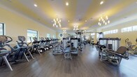 This fitness center, known as the Jim Bissen Center, offers a comprehensive workout facility spread across a single level. The main gym floor is generously sized, featuring warm yellow-striped tray ceilings accented by elegant chandelier lighting and recessed fixtures, creating an inviting atmosphere uncommon in typical fitness spaces. Dark hardwood-style flooring runs throughout, providing a polished, upscale feel. Large windows along the perimeter flood the space with natural light and offer views of the surrounding greenery.

The main floor is outfitted with an extensive row of cardio machines, including treadmills and ellipticals, as well as a full complement of Cybex strength machines, cable systems, free weights, and a dedicated bench press station. WaterRower rowing machines add a premium touch to the cardio offerings.

A dedicated cycling room houses multiple Stages indoor cycling bikes alongside a wall-mounted television, ideal for structured spin sessions. A separate yoga and stretching room provides stability balls, medicine balls, yoga mats, and a flat-screen TV for guided workouts in a calm, uncluttered setting. An entryway area features built-in cubby storage and cabinetry, offering an organized transition space into the facility.
