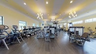 This fitness center, known as the Jim Bissen Center, offers a comprehensive workout facility spread across a single level. The main gym floor is generously sized, featuring warm yellow-striped tray ceilings accented by elegant chandelier lighting and recessed fixtures, creating an inviting atmosphere uncommon in typical fitness spaces. Dark hardwood-style flooring runs throughout, providing a polished, upscale feel. Large windows along the perimeter flood the space with natural light and offer views of the surrounding greenery.

The main floor is outfitted with an extensive row of cardio machines, including treadmills and ellipticals, as well as a full complement of Cybex strength machines, cable systems, free weights, and a dedicated bench press station. WaterRower rowing machines add a premium touch to the cardio offerings.

A dedicated cycling room houses multiple Stages indoor cycling bikes alongside a wall-mounted television, ideal for structured spin sessions. A separate yoga and stretching room provides stability balls, medicine balls, yoga mats, and a flat-screen TV for guided workouts in a calm, uncluttered setting. An entryway area features built-in cubby storage and cabinetry, offering an organized transition space into the facility.