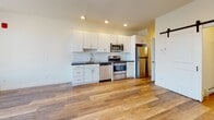 Located at 11 Park St in Essex Junction, Vermont, this single-floor unit offers a thoughtfully designed, open-concept layout with contemporary finishes throughout. Warm wood-style flooring runs continuously across the living and kitchen areas, creating a cohesive and inviting atmosphere.

The kitchen is a standout feature, outfitted with crisp white cabinetry, dark countertops, a subway tile backsplash, and a full suite of stainless steel appliances including a gas range, dishwasher, microwave, and refrigerator. Recessed lighting overhead keeps the space well-lit and functional.

The open living area is bright and airy, benefiting from a large picture window that brings in generous natural light and frames views of the surrounding Essex Junction neighborhood. A wall-mounted mini-split unit provides efficient climate control, complemented by baseboard heating. A stylish sliding barn door adds a modern architectural touch to the interior.

The bathroom is finished with large-format tile flooring, full-height tile in the walk-in glass-enclosed shower, a dark-toned vanity with ample storage, and a white countertop, lending a clean and polished look. Additional storage is accessible via a dedicated closet space off the main living area. The unit's clean, neutral palette and modern fixtures make it a versatile and move-in ready space.