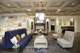 This sophisticated single-floor lounge space showcases a masterful blend of classic architectural details and contemporary interior design. The standout coffered ceiling, finished in warm wood tones with dark accent molding, lends a sense of grandeur and vertical dimension to the room. Dramatic cage-style pendant chandeliers hang throughout, casting warm, layered lighting across the open floor plan.

The herringbone-patterned hardwood flooring grounds the space with timeless elegance, while large multi-pane windows along one wall flood the interior with abundant natural light and frame views of the surrounding exterior. Sheer floor-to-ceiling drapery softens the window walls, adding texture and warmth.

A striking natural stone fireplace serves as a focal point in one seating area, flanked by built-in shelving and a dark navy-painted surround. Adjacent to it, white wainscoting and decorative wall panels add architectural refinement. A curated gallery wall of framed artwork in gold accents adds personality and visual interest throughout the space.

Furnishings are thoughtfully arranged into distinct seating zones, featuring a plush navy tufted sofa, cream wingback chairs, and upholstered accent seating on richly patterned blue and gold area rugs. A bar-height seating area with counter stools completes the layout, making this space ideal for both relaxed gatherings and social interaction.