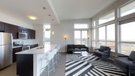 This 2-bedroom, 2-bathroom residence is designed around an open-concept floor plan that seamlessly integrates the kitchen, dining, and living areas into one cohesive space. The kitchen features dark cabinetry contrasted by light countertops, a built-in sink, and a breakfast bar with white modern barstools, illuminated by sleek pendant lighting overhead. The living area is anchored by expansive windows that wrap around the exterior walls, flooding the space with natural light and offering sweeping views of the surrounding landscape and roadways beyond. Elevated ceilings with clean architectural lines enhance the sense of openness throughout. The living room is well-appointed, featuring a leather sofa set arranged around a glass coffee table atop a bold geometric black-and-white area rug. Neutral-toned flooring and white walls create a bright, contemporary backdrop that complements the modern furnishings. The overall ambiance is sleek and sophisticated, characterized by a monochromatic palette, abundant natural light, and thoughtful design details that make efficient use of the single-floor layout.