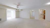 Located at 100 Golden Ln in Dover, Delaware, this single-floor residence offers a clean, open layout designed for comfortable everyday living. The main living area is generously proportioned, featuring plush wall-to-wall carpeting in a warm neutral tone that carries throughout the space. A ceiling fan is centrally positioned overhead, providing both comfort and airflow. Large vertical blinds line the windows along one wall, allowing for adjustable natural light while maintaining privacy. The walls are finished in a crisp, bright white, creating a clean and airy atmosphere that enhances the sense of openness. A hallway extends from the main living area, leading toward additional rooms and a paneled entry door visible at the far end, suggesting a well-organized interior layout. The overall ambiance of the space is calm and understated, with a neutral palette that provides a versatile backdrop suited to a variety of interior styles. The single-floor design ensures seamless accessibility and ease of movement throughout. This residence at 100 Golden Ln presents a straightforward, functional living environment in Dover, DE.