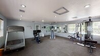 Located at 726 North McDonald Road in Spokane, Washington, this fitness center offers a well-equipped, single-floor exercise facility designed to support a variety of workout routines. The space is finished with neutral gray carpeting throughout and light blue-gray painted walls, creating a clean and functional environment.

The main workout area is generously sized and features an array of cardio equipment, including a treadmill, stationary bike, stair climber, and elliptical machine. A multi-station cable weight machine and a weight bench provide dedicated strength training options. A large wall-mounted mirror along one side of the room visually expands the space and allows users to monitor their form during exercise.

Natural light filters in through multiple windows fitted with blinds, offering views of the surrounding greenery. Recessed ceiling lighting supplements the daylight, ensuring the room remains well-lit throughout the day. A textured ceiling adds a subtle architectural detail overhead.

Near the entrance, a wall-mounted television and a wall clock provide practical amenities for users during their workouts. A wooden bench near the entry area offers a convenient spot for changing footwear, while wall-mounted hooks provide storage for personal items. A glass-paned entry door connects the fitness room to the broader property, completing this functional and accessible community amenity.