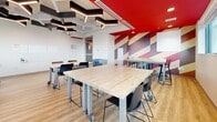 Located at 22 Bay St S in Hamilton, Ontario, this dynamic single-room collaborative workspace delivers a modern and energetic environment designed to inspire productivity and creativity. The room is anchored by light wood-finish tables on casters, offering flexible reconfiguration to suit a variety of group sizes and working styles, complemented by sleek black stackable chairs.

The ceiling is a standout architectural feature, showcasing striking geometric light fixtures in hexagonal and Y-shaped forms, interspersed with acoustic panels that help manage sound in the open layout. A bold red ceiling panel adds a vivid pop of colour, reinforced by a dramatic diagonal graphic wall treatment in red, cream, and burgundy tones that energizes the space.

Large horizontal windows wrap along one wall, flooding the room with natural light and offering sweeping views of the Hamilton cityscape, providing an inspiring urban backdrop. Light wood-plank flooring grounds the space warmly and contrasts effectively with the high-contrast décor.

A full-width whiteboard is mounted along one wall, supporting presentations and collaborative brainstorming. Recycling stations near the entrance reflect a commitment to sustainability. A glazed door provides visibility and connectivity to adjacent areas. This thoughtfully designed space is well-suited for workshops, team meetings, and collaborative work sessions.