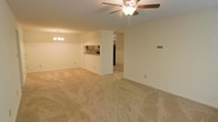 This single-floor space features an open-concept layout that seamlessly connects the living, dining, and kitchen areas into one expansive, flowing floor plan. The living area is finished with wall-to-wall beige carpeting, lending a warm and neutral aesthetic throughout. A ceiling fan with a built-in light fixture is centrally mounted overhead, providing both air circulation and ambient lighting to the main living space.

Adjacent to the living area, a decorative chandelier marks the dining zone, adding a subtle elegance to the open layout. The kitchen is partially separated by a structural half-wall featuring a pass-through counter, which maintains visual connectivity while defining the cooking space. Wooden cabinetry and a dark countertop are visible through the pass-through, along with what appears to be a microwave mounted above the counter.

The walls throughout are finished in a soft cream tone, contributing to the bright and airy feel of the interior. A doorway visible beyond the kitchen suggests access to additional rooms or utility areas. The overall ambiance is clean, understated, and functional, well-suited for everyday living.