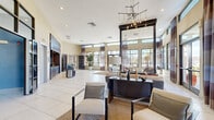 This generously proportioned single-floor space showcases a sophisticated contemporary design with an open, airy layout that maximizes both functionality and visual appeal. The main living area is defined by soaring double-height ceilings that create an impressive sense of volume, complemented by an abundance of natural light streaming through floor-to-ceiling windows and transom windows along the perimeter walls. Dark-framed window systems are dressed with layered drape panels in warm earth tones, adding texture and dimension to the space.

A dramatic focal point is provided by a large-scale mosaic accent wall with an organic, cellular pattern, further elevated by an angular, industrial-style chandelier suspended above. Large-format white tile flooring runs throughout, lending a clean, cohesive foundation to the design. Distinct seating zones are defined by patterned area rugs and contemporary upholstered furniture in neutral tones.

A built-in wet bar or service counter with bar seating adds a functional amenity, while tall interior doors with glass insets provide connectivity to adjacent rooms, including a private office area with bold blue accent walls and a textured wall-mounted media unit. Potted indoor trees and curated décor complete the polished, upscale ambiance.