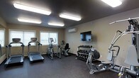 This fitness room is designed to provide a functional and comfortable workout environment. The space is finished with dark, rubberized flooring that offers durability and safety underfoot. Neutral beige walls and a dark ceiling create a focused atmosphere, while flush-mounted fluorescent light fixtures provide consistent, bright illumination throughout.

A row of windows along one wall allows natural light to filter in, adding an open feel to the space. A wall-mounted mini-split HVAC unit ensures climate control year-round, keeping the environment comfortable during any season.

The room is equipped with a variety of cardio machines, including a treadmill, an upright exercise bike, and an elliptical trainer or cross-trainer, offering users a range of cardiovascular workout options. A multi-tiered dumbbell rack stocked with a full range of free weights supports strength training needs. A flat-screen television is mounted on the wall, allowing users to stay entertained during workouts. Additional equipment is partially visible, suggesting the space is comprehensively outfitted for a well-rounded fitness routine.
