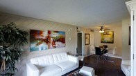 This single-floor space showcases a thoughtfully designed open-concept layout that seamlessly connects the living and dining areas. The living room is anchored by a sleek white tufted sofa and matching ottoman, resting atop a patterned area rug over rich dark hardwood flooring. A striking diagonal shiplap wood-paneled accent wall serves as a bold architectural focal point, lending texture and character to the space. A large, vibrant abstract canvas painting in red, blue, and yellow tones adorns this wall, adding an artistic and energetic element to the room.

The open sightline leads directly into the dining area, where a ceiling fan with integrated lighting provides both function and style. Dark modern dining chairs surround a compact table, creating an intimate setting ideal for meals. Framed artwork decorates the dining area wall, complementing the overall aesthetic.

A doorway visible from the living area hints at additional rooms beyond, including what appears to be a closet. The textured ceiling and visible HVAC vents indicate practical, well-considered infrastructure throughout. The overall ambiance is modern and artistic, with a clean, contemporary feel.