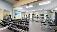 Located at Charter Creek in Ashland, Virginia, this fitness center provides a comprehensive workout facility spread across a single, generously sized floor. The space is organized into distinct zones designed to support a full range of exercise routines. The main cardio area features multiple treadmills, each equipped with individual television screens, alongside elliptical machines and steppers—all positioned in front of large wall mirrors that visually expand the space and allow users to monitor their form. Natural light filters in through numerous windows fitted with white shutters, creating a bright and energizing atmosphere enhanced by ceiling fans and recessed fluorescent lighting overhead. A dedicated free weights section houses a full rack of dumbbells in graduated sizes, accompanied by workout benches and stability balls in multiple sizes stored on a vertical rack. A separate strength-training area contains a multi-station cable and weight machine, an additional elliptical, and open floor space for functional training. The flooring throughout is durable rubber, ideal for high-traffic athletic use. The interior walls are painted in a warm cream and accented with bold teal geometric designs, giving the facility a modern and motivating aesthetic. An entry area with a branded Charter Creek mat and a small bookshelf rounds out the space, offering a welcoming transition into the fitness floor.