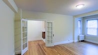 This single-floor space offers a clean, well-maintained interior defined by classic architectural details and a warm, neutral aesthetic. The main living area is finished with beautiful light hardwood floors that run throughout, lending a sense of warmth and continuity to the space. The walls are painted in a soft, off-white tone that enhances the bright and airy ambiance of the interior.

A standout feature of the space is the pair of elegant French doors with multi-pane glass panels, which divide the rooms while allowing natural light to flow freely between them. These doors add a charming, classic character to the layout and create a sense of openness without sacrificing definition between spaces.

Beyond the French doors, a connected room is visible, offering a glimpse of additional living or functional space. A small wooden cabinet or sideboard can be seen in the background, hinting at the utility and versatility of the adjoining area.

White baseboard trim runs along the walls throughout, complementing the light hardwood floors and contributing to the crisp, cohesive finish of the interior. The layout is straightforward and functional, making it an adaptable space suitable for a variety of uses. Overall, the space combines timeless design elements with a bright, inviting atmosphere.