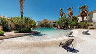 Located at 435 Ithaca Ct in Las Cruces, New Mexico, this beautifully designed outdoor community space exudes a resort-like atmosphere under the bright New Mexico sky. The centerpiece is a generously sized freeform swimming pool with crystal-clear turquoise water, complemented by a distinctive beach-entry section with a sandy shoreline perfect for relaxed lounging. Surrounding the pool are multiple patio areas furnished with chaise lounges, offering ample space for sunbathing and outdoor relaxation.

The landscaping is lush and thoughtfully designed, featuring towering palm trees, mature shade trees, ornamental shrubs, and desert-adapted plantings that create a private, oasis-like environment. Stone masonry walls and pillars — constructed from natural stacked rock — define the perimeter and gated entry, lending a high-end, rustic elegance to the space.

A covered pavilion with a tiled Spanish-style roof serves as a social hub, housing a bar-height counter and seating, as well as restroom facilities. Winding pathways meander through the landscaped grounds, connecting the various areas of the property. The main building, featuring a red-tile roof, stucco exterior, and arched architectural details consistent with Southwest Mediterranean design, overlooks the pool and grounds, completing the cohesive and inviting aesthetic of this exceptional outdoor space.