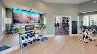 This dedicated fitness and yoga room offers a versatile, well-appointed space designed to support a wide range of workout routines. The room features light hardwood-style flooring throughout, providing a clean, durable surface ideal for both equipment-based and floor exercises. Muted sage-green walls create a calm, focused atmosphere, while large windows fitted with white blinds line multiple walls, flooding the space with abundant natural light and offering views of lush greenery outside. A full-length wall mirror expands the sense of space and allows for form-checking during workouts. The room is comprehensively equipped with a row of spin bikes for cardio training, a rack of color-coded dumbbells and kettlebells, a BOSU balance trainer, medicine balls, stability balls, and resistance equipment neatly organized on a freestanding rack. A large wall-mounted display screen with an interactive fitness platform enables guided workout sessions. A wall-mounted fold-down mat or bench adds additional versatility. An open doorway connects directly to an adjacent fitness area, making this studio part of a broader, cohesive fitness facility. Recessed lighting supplements the natural light, ensuring the space remains bright and functional at all hours.