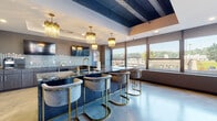 Located at 901 Lee St in Charleston, West Virginia, this stylish amenity floor delivers a refined, modern atmosphere designed for socializing and relaxation. The open-plan layout flows seamlessly between distinct zones, all united by polished concrete flooring and an elevated design aesthetic.

The central bar and kitchen area serves as the heart of the space, anchored by a dramatic dark granite island with gold-accented bar stools and illuminated by cascading crystal-and-brass chandeliers. Hexagonal tile backsplashes, dark wood cabinetry, a beverage refrigerator, and dual wall-mounted televisions complete this well-appointed entertaining hub.

A billiards zone features a statement pool table positioned beneath exposed dark structural beams and soaring ceilings, flanked by plush seating arrangements and floor-to-ceiling windows that frame sweeping views of the Charleston skyline.

The lounge area offers a warm contrast with rich burnt-orange velvet sofas arranged around a geometric coffee table, set against a striking mixed-media accent wall that adds artistic depth and texture to the room. Rustic wood floating shelves provide an earthy balance to the contemporary palette.

Throughout the space, expansive windows flood the interiors with natural light and provide elevated views of the surrounding cityscape. A secondary kitchenette with granite countertops and a coffee station rounds out the functional offerings of this thoughtfully designed communal retreat.