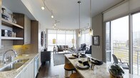 This single-floor space showcases a thoughtfully designed open-concept layout that seamlessly connects the kitchen, dining, and living areas into one cohesive environment. The kitchen features granite countertops and a large island with bar seating, illuminated by a series of elegant pendant lights that draw the eye toward the dining area. Track lighting along the ceiling provides additional flexibility in highlighting key areas throughout the space.

The living area is anchored by a generous seating arrangement and benefits from an abundance of natural light streaming through expansive floor-to-ceiling windows. These windows wrap around the corner of the space, offering sweeping city views and creating a bright, airy atmosphere. Dark hardwood flooring runs throughout, adding warmth and contrast against the neutral-toned walls and light ceilings.

The overall ambiance is contemporary and refined, with a carefully curated blend of modern furnishings, layered lighting, and high-end finishes. The open sightlines between functional zones create a sense of spaciousness, making the layout well-suited for both everyday living and entertaining.