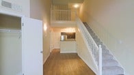 This two-story residence offers a thoughtfully designed layout that maximizes both space and functionality. The main level immediately impresses with its dramatic double-height ceiling, which creates a sense of openness and airiness throughout the living area. Rich hardwood flooring extends across the ground floor, lending warmth and a polished aesthetic to the space.

The kitchen is positioned at the rear of the main level and features dark cabinetry paired with stainless steel appliances, including a refrigerator, and a breakfast bar-style counter that provides both workspace and a casual dining option. Overhead track lighting illuminates the kitchen area effectively.

A classic white-baluster staircase with a carpeted runner leads to the upper level, where an open loft overlooks the main living area below. The white painted railings of the upper-floor balcony add a clean, traditional touch that contrasts nicely with the warm tones of the lower level.

Additional doors visible on the main floor suggest access to closets or utility spaces, contributing to the overall practicality of the layout. Wall-mounted sconce lighting adds a soft, welcoming ambiance. The combination of soaring ceilings, open sightlines between floors, and cohesive neutral tones gives this residence a comfortable yet spacious character.