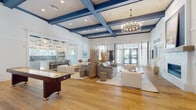 Located at 205 Hunters Lake Way in Ponte Vedra, Florida, this single-story community space embodies a refined coastal aesthetic throughout its generously proportioned interior. The main gathering area immediately impresses with soaring ceilings adorned with bold navy blue coffered beams, which contrast beautifully against crisp white walls and warm hardwood flooring. A statement chandelier anchors the space from above, while recessed lighting adds a soft, layered glow throughout.

The lounge area is thoughtfully arranged with neutral-toned upholstered seating, area rugs, and organic decorative elements such as tall driftwood arrangements and ceramic vessels, reinforcing the coastal atmosphere. A sleek linear fireplace set into a white tile surround serves as a focal point along one wall, complemented by large French doors that invite natural light and offer views to the outdoors.

A game area with a foosball table provides recreational variety within the open floor plan. Adjacent to the main lounge, a well-appointed kitchen features white shaker cabinetry, quartz countertops, stainless steel appliances, and a striking blue penny-tile backsplash. Bamboo Roman shades filter natural light while maintaining the relaxed, beachy character that defines this welcoming community gathering space.