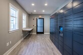 Located at 8064 S Fulton Pkwy in South Fulton, Georgia, this dedicated package room is a well-appointed, single-floor amenity space designed to streamline parcel management. The room features light wood-look luxury vinyl plank flooring throughout, paired with neutral gray walls and crisp white baseboards and window trim, creating a clean and modern aesthetic.

A prominent wall of Amazon Hub smart lockers lines one side of the room, offering secure, automated package retrieval for residents. These are complemented by additional large-format parcel lockers on the adjacent wall, accommodating oversized deliveries. A built-in countertop shelf along one wall provides a convenient surface for sorting and staging packages.

Natural light enters through multiple white-trimmed windows, offering views of the surrounding exterior grounds and keeping the space bright and inviting. Recessed ceiling lighting ensures consistent illumination throughout all hours. A glass-paneled exterior access door provides direct entry for delivery personnel, while safety features including illuminated exit signage and fire safety hardware are clearly visible.

The overall layout is open and functional, offering ample floor space to comfortably move and manage deliveries. This thoughtfully designed package room reflects a commitment to modern convenience and practical, resident-focused amenities.