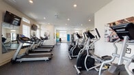 Located at 1000 Kenmore Crossing Blvd. in Marysville, Ohio, this well-appointed fitness center offers a spacious, single-floor layout designed to support a full range of workout routines. The facility is bathed in natural light through large windows fitted with white shutters and transom windows above, creating an open and energizing atmosphere. Recessed lighting and ceiling fans throughout ensure consistent illumination and airflow. The durable speckled rubber flooring provides both safety and practicality across the entire space. The cardio zone is well-equipped with multiple treadmills and elliptical machines, each paired with individual display screens, and wall-mounted flat-screen TVs for entertainment during workouts. A dedicated strength training area features a multi-functional cable machine, a rack of dumbbells, an adjustable weight bench, and additional resistance equipment. Full-length mirrors line portions of the walls, enhancing the sense of space while supporting proper form. Exercise accessories, including stability balls and yoga mats, are also available. Windows overlooking what appears to be an outdoor pool area add a pleasant visual connection to the broader amenity grounds. The facility also includes a separate restroom accessible from within the space.