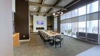Located at 23210–23230 Chagrin Blvd in Beachwood, Ohio, this single-floor community gathering space combines contemporary design with warm architectural details to create an inviting, functional environment. The room's most distinctive overhead feature is its coffered ceiling with exposed rustic wood beams framing recessed panels, lending a refined yet organic character to the space. Pendant lighting with a modern, sculptural design hangs above the central area, complementing the recessed can lighting integrated throughout the ceiling.

A generously sized rectangular dining table with upholstered side chairs serves as the focal point of the room, accommodating a large number of guests and making the space well-suited for gatherings and events. Along one wall, a sleek linear fireplace anchors a clean white accent wall, above which a wall-mounted flat-screen television is positioned for easy viewing. Decorative accents are thoughtfully placed throughout, adding visual interest without cluttering the space.

Floor-to-ceiling windows along one side of the room flood the interior with natural light and provide direct views of and access to an adjacent outdoor patio area, seamlessly connecting indoor and outdoor living. The stone-tile flooring adds texture and durability, grounding the overall aesthetic in a sophisticated, contemporary style.