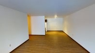 This single-floor space presents a clean, open layout designed for practical living. The main living area is generously proportioned, featuring warm hardwood-style flooring that runs throughout, lending a cohesive and inviting feel to the space. Crisp white walls reflect light effectively, contributing to a bright and airy atmosphere despite the absence of prominent windows in the main area.

A half-wall partition subtly divides the living space, adding a sense of architectural definition without compromising the overall openness of the floor plan. Beyond the partition, the space transitions into a kitchen area, which is partially visible and equipped with standard appliances including a full-size refrigerator. The kitchen is tucked neatly into its own designated zone, keeping it functional yet integrated with the broader living space.

Overhead lighting in the kitchen area provides focused illumination, while the smooth, white ceiling throughout maintains a clean and uncluttered aesthetic. Dark baseboards offer a subtle contrast against both the white walls and the warm flooring, adding a finishing detail that ties the interior together.

Overall, this single-floor space offers a straightforward, well-maintained layout that balances open living with defined functional zones, making it a versatile and efficient environment.