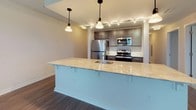 This well-appointed 3-bedroom, 2-bathroom single-floor residence offers a thoughtfully designed open-concept layout that seamlessly connects the main living areas. Rich, dark hardwood flooring runs throughout the living and dining spaces, complementing the neutral wall tones and clean white trim that create a bright, welcoming atmosphere. Multiple windows allow generous natural light to fill the living room, while a glass-panel door opens directly onto a private balcony with outdoor views.

The kitchen serves as a central focal point, featuring a large island with a light granite countertop, an undermount sink, and stylish pendant lighting overhead. Taupe cabinetry pairs with stainless steel appliances — including a refrigerator, range, and built-in microwave — and track lighting illuminates the workspace efficiently.

Each of the three bedrooms is finished with plush carpeting, closets, and neutral walls that offer a calm, restful environment. One bedroom benefits from a large window with outdoor views, adding to the sense of openness.

Both bathrooms are finished with large-format tile flooring, dark vanity cabinetry with stone-topped counters, and full tub-shower combinations with chrome fixtures, providing a clean and functional design throughout.