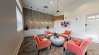 Located at 103 Inspiration Ave in Bluffton, SC, this single-floor community amenity space blends coastal charm with modern functionality. The open layout is anchored by a central lounge area furnished with a set of vibrant coral-upholstered accent chairs arranged around a sculptural round coffee table, creating an inviting gathering spot. A striking rustic wood pendant light hangs overhead, complementing the space's warm yet refined aesthetic.

Crown molding and crisp white walls provide a clean architectural backdrop, while patterned carpet in neutral tones grounds the room with visual texture. One wall features a beadboard-paneled accent section painted in a soft aqua hue, adorned with framed botanical and coral-inspired artwork that reinforces the coastal theme. Bold abstract paintings on adjacent walls add an artistic contrast.

Along one wall, a built-in business center offers multiple computer workstations and ergonomic seating, making the space well-suited for productivity. Large windows with white blinds flood the area with natural light and offer views of the surrounding greenery.

A wet bar area along the opposite wall is finished with white cabinetry, a sleek marble-look countertop, a statement matte black faucet, and floor-to-ceiling large-format tile, adding a sophisticated, upscale touch to this well-appointed community space.