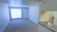 This single-floor space features an open-concept layout that connects the living area to a partial kitchen counter, creating a practical and cohesive flow. The living area is finished with wall-to-wall carpet in a neutral gray tone, providing a soft underfoot feel throughout the space. A large window along one wall allows natural light to fill the room, contributing to a bright and airy atmosphere. The front entry door is positioned adjacent to the window, offering a straightforward and accessible entryway. Walls are painted in a soft, neutral tone that complements the overall understated aesthetic. The textured ceiling adds a subtle design element consistent with the unit's functional style. On the kitchen side, a granite-topped counter extends into the space, hinting at a partially open kitchen layout separated by a half-wall partition. A smoke detector is visibly mounted near the ceiling, and standard electrical outlets are positioned throughout the walls. The space presents a clean, move-in-ready condition with a modest and practical design suited for everyday living.