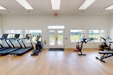 This fitness center in Valdosta, Georgia offers a bright, modern exercise space designed to support a full range of workout activities. The room features soaring ceilings with recessed skylight panels that flood the interior with natural light, complemented by large windows along the walls that frame outdoor views of the surrounding greenery. Light hardwood flooring runs throughout, creating a clean and inviting atmosphere.

The space is thoughtfully equipped with a variety of cardio machines, including treadmills, an elliptical, and a stationary cycling bike, all positioned to take advantage of the natural light and outdoor views. A multi-station cable machine provides options for comprehensive strength training, while a dedicated free weights area includes a full dumbbell rack and an adjustable workout bench. A medicine ball and stability ball rack adds versatility for functional fitness routines.

A small beverage refrigerator offers added convenience for hydration during workouts. A water fountain is also available within the space. Double glass doors on both ends of the room provide easy access and allow for excellent cross-ventilation. The crisp white walls and open floor plan create a spacious, uncluttered environment well-suited for individual workouts or small group fitness sessions.
