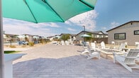 Located at 4747 N 99th Ave in Phoenix, Arizona, this single-level outdoor community amenity space is thoughtfully designed to complement the desert Southwest lifestyle. The centerpiece is a generously sized paver patio finished in warm, neutral tones, providing ample room for social gatherings and relaxation. White Adirondack chairs and umbrella-shaded seating clusters are spread throughout the space, encouraging outdoor enjoyment under the Arizona sky.

A covered outdoor lounge area features exposed dark wood beam ceilings, ceiling fans, large glass doors, and a striking whitewashed brick fireplace, creating a comfortable retreat that bridges indoor and outdoor living. Adjacent to the lounge is a built-in outdoor grill station, ideal for entertaining.

A fenced swimming pool is visible along the perimeter, accompanied by desert-appropriate landscaping including gravel beds, native shrubs, and young palms. A dedicated green lawn area provides space for lawn games such as cornhole, adding a recreational dimension to the community.

The overall ambiance is clean, modern, and resort-inspired, with consistent use of teal market umbrellas, white furniture, and stone-accented architectural details tying the various zones together. The open layout allows for clear sightlines across the community, making this a welcoming and functional shared outdoor environment.