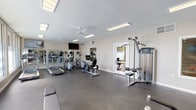 Located at 1700 Via Pacifica in Corona, California, this single-floor fitness center is a well-equipped facility designed to support a comprehensive workout experience. The space is organized across two distinct zones, each thoughtfully appointed for different types of exercise.

The main workout area features durable rubber flooring and is outfitted with a variety of cardio machines, including treadmills, ellipticals, and a stationary bike. A cable resistance machine provides strength-training options, while a flat utility bench and a rack stocked with medicine balls, stability balls, and dumbbells cater to functional and free-weight training. Full-length mirrors along one wall expand the sense of space and allow for form monitoring, while ceiling fans and recessed panel lighting keep the environment comfortable and well-lit. A wall-mounted television adds an entertainment element for cardio sessions.

A secondary room features light wood-style flooring, recessed can lighting, and a dedicated cycling area equipped with multiple Life Cycle upright bikes and a spin bike, creating an ideal environment for low-impact cardio training. A wall-mounted air conditioning unit ensures climate control in this space.

Large windows in the free weights area allow natural light to filter in, offering views of the surrounding greenery and contributing to an open, energizing atmosphere throughout the facility.