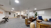 Located at 2450 Island Dr in Spring Park, MN, this single-floor community amenity space offers a generously sized, open layout designed for both socializing and relaxation. The main living area features patterned carpet tiles throughout, white shiplap wainscoting along the lower walls, and recessed lighting that keeps the space bright and welcoming. A nautical theme is carried throughout, with life ring and anchor décor accents adding a charming lakeside character.

The living area is divided into distinct functional zones. A lounge area provides comfortable seating, including tufted blue wingback chairs, a lattice-back armchair, and a wall-mounted flat-screen TV — ideal for casual gatherings. A game zone features a shuffleboard table and a high-top bistro table with bar stools for recreational use. Toward the back, a kitchenette with bar seating, pendant lighting, and hardwood-style flooring serves as a hub for entertaining.

A dining area with a large table and ample seating accommodates group meals or meetings, while a structural column adds an architectural element to the open floor plan. A separate utility area includes a full-size refrigerator for additional storage. The space also includes a dedicated laundry room and coat hooks for added convenience, making this a fully functional and thoughtfully designed communal retreat.