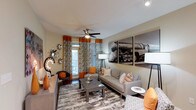 This single-floor, one-bedroom, one-bathroom residence offers a thoughtfully designed living space with a distinctly modern aesthetic. The living room serves as the focal point of the home, showcasing hardwood-style flooring topped with a patterned area rug that anchors the seating arrangement. A gray sofa and coordinating accent chair are styled with orange and patterned throw pillows, creating a warm, cohesive color palette carried throughout the room via bold orange curtains and decorative wall accents. A ceiling fan with integrated lighting provides both airflow and ambient illumination, complementing the floor lamps positioned throughout the space. A large black-and-white photographic print of a vintage locomotive commands attention as a dramatic wall feature, adding an industrial artistic element to the interior. A glass-top coffee table and side table complete the living area furnishings. A sliding glass door or large window allows natural light to filter in, brightening the space and providing a connection to the outdoors. The overall ambiance is modern and energetic, with carefully layered décor that balances bold visual interest with comfortable, functional living.