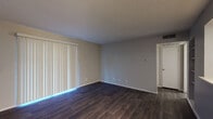 This single-story space offers a functional and straightforward layout designed for comfortable everyday living. The main living area is finished with dark hardwood-style flooring that extends throughout, complemented by neutral gray walls and a textured ceiling. A large sliding glass door fitted with vertical blinds allows natural light to fill the room while providing access to the exterior. A pass-through counter connects the living area to the kitchen, creating an open, communicative flow between the two spaces. Built-in shelving adjacent to the pass-through adds practical storage.

The kitchen is equipped with warm wood-toned cabinetry, light countertops, and black appliances including a refrigerator and electric range. Track lighting overhead provides focused illumination throughout the cooking area.

The bedrooms are carpeted for comfort and feature windows with horizontal blinds that allow natural light. One bedroom includes a ceiling fan with integrated lighting. The bathroom offers a wide vanity with ample counter space, a large mirror with overhead lighting, and dark hardwood-style flooring that mirrors the living areas. A combination tub and shower completes the bathroom's practical design. Throughout the unit, white baseboards provide a clean contrast against the flooring and wall tones.