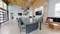 Located at 3 Woodcock Rd in Savannah, Georgia, this single-floor community gathering space combines industrial-modern design with warm natural materials to create an inviting and versatile environment. The centerpiece of the room is a striking oversized granite-topped island flanked by teal upholstered counter stools, illuminated from above by a row of matte black pendant lights. The soaring ceiling features exposed natural wood planking accented by timber cross-beams and industrial-style ductwork, lending the space a refined yet relaxed loft character. Polished concrete tile flooring extends throughout, tying together the open floor plan seamlessly. A built-in wet bar lines one wall, complete with white cabinetry, a stainless steel sink, a beverage refrigerator, and a large wall-mounted television — ideal for gatherings and events. Multiple lounge seating areas are arranged throughout, including tan leather benches with coordinating ottomans and coral-toned accent chairs, each positioned near wall-mounted screens. Black-framed glass garage-style doors along one wall flood the interior with natural light while connecting the space visually to the outdoors. A billiards table rounds out the entertainment offerings. The overall ambiance is contemporary, airy, and well-appointed, making this a dynamic and functional communal space.