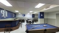 This single-floor recreation space is fully dedicated to Memphis Grizzlies fandom, featuring team branding, logos, and a navy blue and gray color scheme throughout. The open layout is designed for entertainment and social interaction, with several distinct zones that flow seamlessly into one another.

At the forefront is a full-size pool table dressed in team-branded navy blue felt, complete with cues and balls ready for play. Adjacent to the pool table is a wooden bar counter with bar stools, providing a casual spot for refreshments and conversation. The bar area is accented with Grizzlies signage, reinforcing the team-centric aesthetic.

The lounge area at the rear of the space features multiple seating arrangements, including round tables with chairs, well-suited for group gatherings or casual dining. Structural columns in the space are wrapped in matching navy blue, contributing to the cohesive design.

The walls are adorned with prominent Memphis Grizzlies bear logos and team emblems, creating an immersive fan experience. Overhead, a drop ceiling with recessed panel lighting provides bright, even illumination throughout the space. The light tile flooring is clean and durable, complementing the overall layout. This space serves as a compelling venue for fans and groups looking to enjoy leisure activities in a spirited, team-inspired environment.
