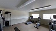 This single-floor fitness room offers a clean, functional environment designed for cardio and light exercise. The space features neutral beige walls paired with carpeted flooring, creating a comfortable setting for workouts. Current equipment includes a treadmill and a stationary upright bike, along with a workout bench, providing a solid foundation for cardiovascular and strength training routines.

Natural light enters through two windows positioned along the far wall, keeping the room bright and airy. Overhead, linear fluorescent lighting supplements natural illumination, ensuring consistent visibility throughout the day. The ceiling features subtle structural beams that add a modest architectural detail to the otherwise straightforward design.

The room's layout allows for adequate spacing between equipment, promoting ease of movement and safe use of each machine. A chair rail–style accent line runs along the lower portion of the walls, adding a simple decorative touch. Electrical outlets are conveniently located along the walls, allowing for additional equipment or accessories. Overall, the space provides a practical, no-frills fitness environment suited for everyday exercise needs.