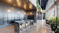 Located at 2990 Longwater Blvd in Kissimmee, Florida, this contemporary residential lobby delivers a sophisticated first impression through a carefully curated blend of industrial and modern design elements. The space is anchored by a dramatic black marble island with bold white veining, surrounded by sleek bar-height seating — an ideal spot for residents to sort mail or gather briefly. Polished concrete floors stretch throughout, reflecting both natural and ambient light to amplify the sense of openness.

One wall is dedicated to an extensive bank of individual mailboxes alongside a clearly designated Package Room, providing residents with a convenient and organized mail management system. The dark tile cladding on these walls creates a striking contrast against the lighter tones of the surrounding space.

Floor-to-ceiling windows along one side of the lobby flood the interior with abundant natural light while offering views of the lush Florida landscaping outside, including mature palms and well-maintained greenery. Large potted tropical plants are thoughtfully placed throughout, reinforcing a biophilic design aesthetic. Woven pendant light fixtures and trailing vines suspended from above add warmth and texture. The exposed concrete ceiling with visible ductwork lends an industrial edge, while leather seating areas provide comfortable, welcoming nooks for residents.