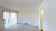 Located at 700 Saint Marys Ave in Frankfort, Indiana, this single-floor unit offers a clean, functional layout designed for comfortable everyday living. The main living area is finished with wall-to-wall carpeting in a neutral tone, providing a soft and welcoming atmosphere throughout the space. Crisp white walls keep the interior feeling bright and open, while a textured ceiling adds a subtle architectural detail overhead. A sliding glass door fitted with vertical blinds lines one wall of the living area, allowing an abundance of natural light to fill the room and providing convenient access to the outdoors. A baseboard heating unit runs along the base of the main wall, ensuring efficient warmth throughout the colder months. A doorway on the adjacent wall connects to the adjoining room, allowing for a practical flow between spaces. The overall design is straightforward and versatile, offering a neutral palette that serves as an ideal canvas for personalization. The single-floor layout ensures easy accessibility throughout, making the space well-suited for a wide range of occupants.